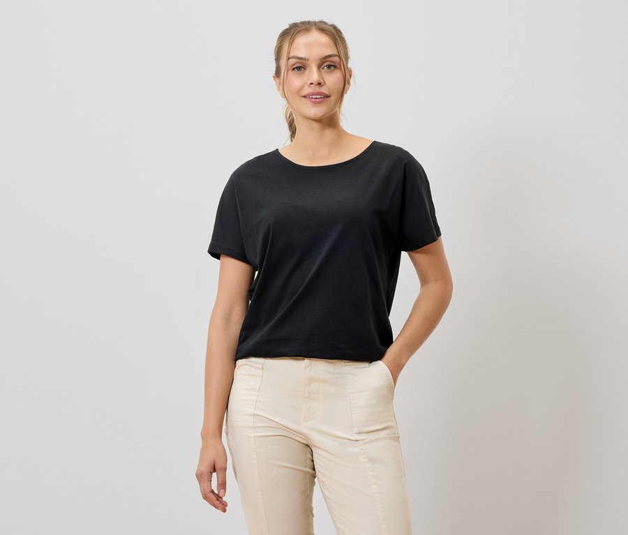 Femme portant un t-shirt noir et un pantalon beige souriant à la caméra.