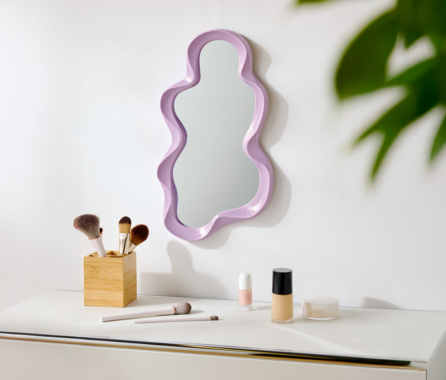 Un miroir violet de forme irrégulière est accroché à un mur blanc au-dessus d'une table blanche avec des pinceaux de maquillage et des cosmétiques.