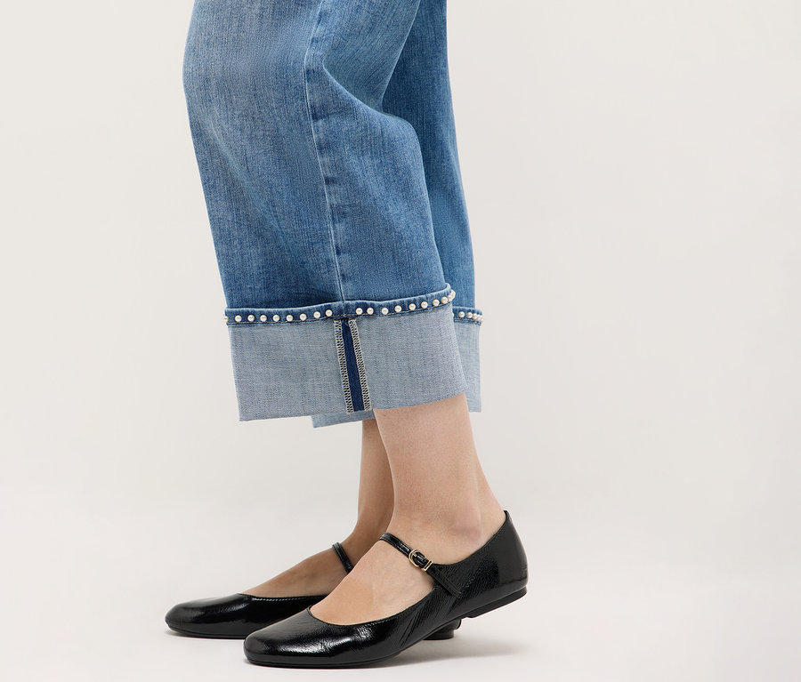Gros plan sur des jambes portant un jean Angels Denim « Linn Cool » associé à des chaussures Mary Jane noires sur un fond neutre.