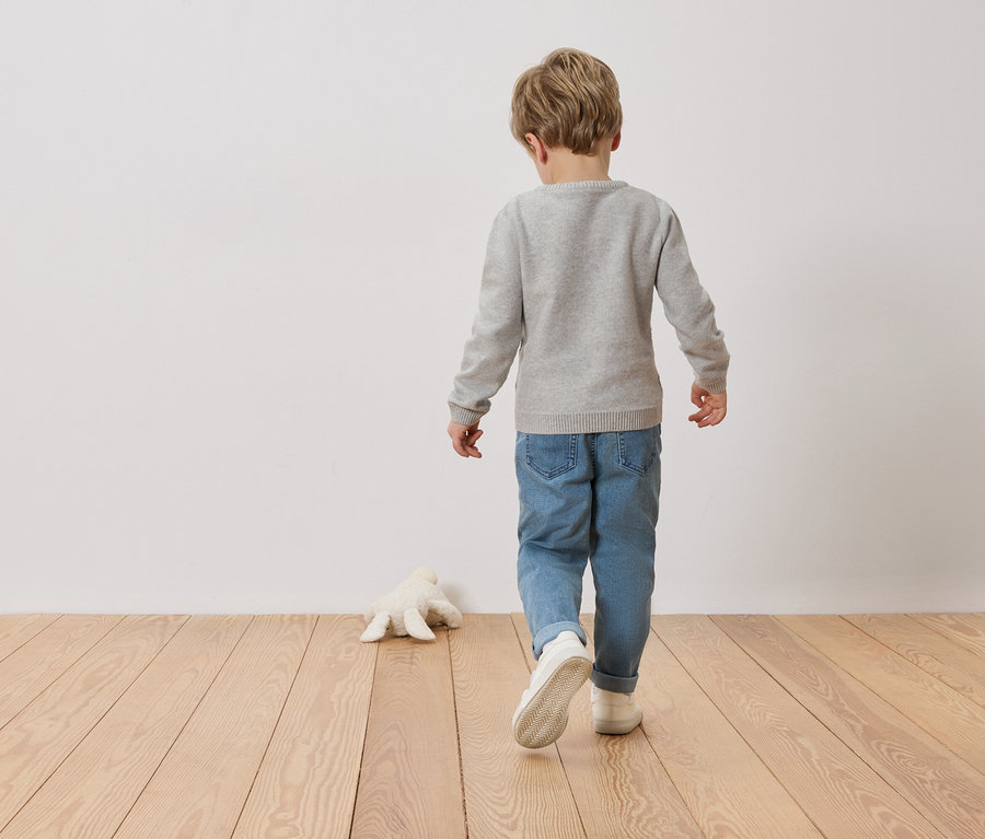 Enfant vu de dos portant un pull gris et un jean, marchant sur un plancher en bois le long d'un mur. Une peluche est posée sur le sol.