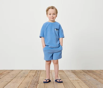 Un garçon se tient devant un mur blanc, vêtu d'un t-shirt bleu avec un motif de requin, d'un short bleu et de tongs bleues.