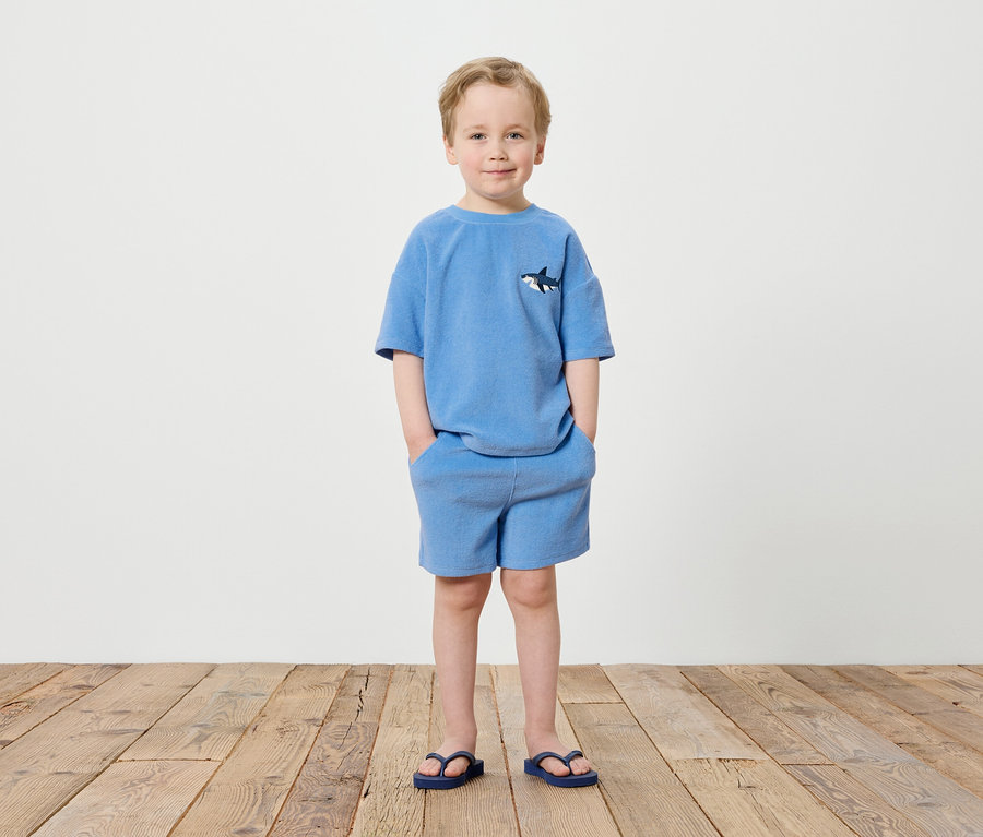 Un garçon se tient devant un mur blanc, vêtu d'un t-shirt bleu avec un motif de requin, d'un short bleu et de tongs bleues.