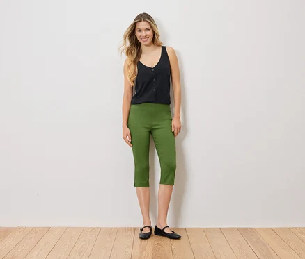 Une femme se tient devant un mur blanc, vêtue d'un haut noir, d'un pantalon Capri vert et de ballerines noires.