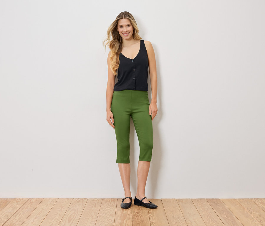 Une femme se tient devant un mur blanc, vêtue d'un haut noir, d'un pantalon Capri vert et de ballerines noires.