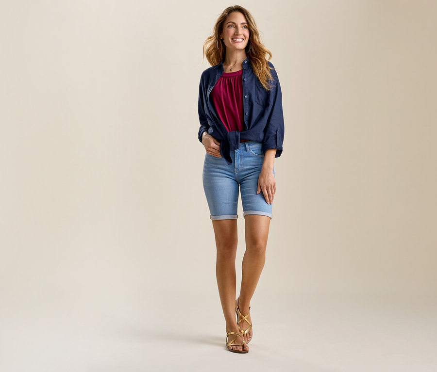 Femme portant un jeans bermuda – coupe « Lea », light blue et des sandales. Elle porte un chemisier bordeaux sous une chemise bleu marine ouverte et nouée.