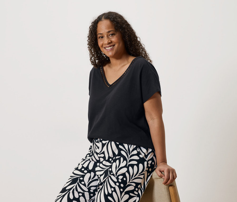 Une femme souriante aux cheveux bouclés, portant un t-shirt noir à col en V et un pantalon à motifs, assise sur une chaise.