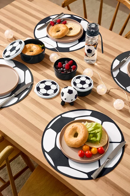 Sur une table en bois sont disposés quatre sets de table, une gourde, trois boîtes à lunch, un ballon de foot en peluche avec son et une guirlande lumineuse à LED « Football ».