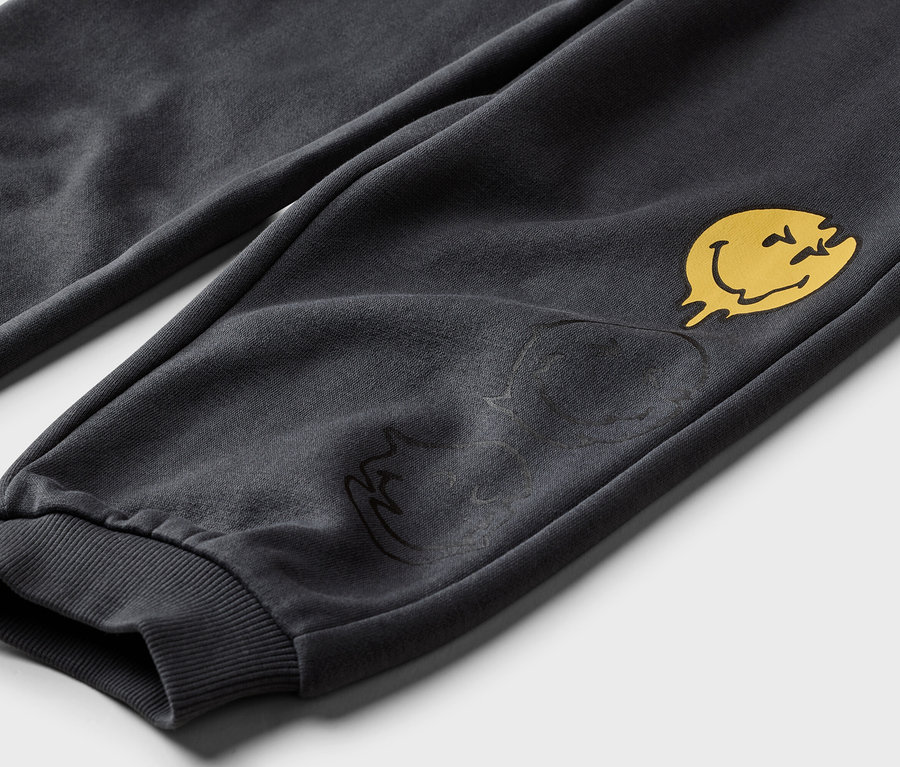 Gros plan d'un pantalon de jogging gris avec un smiley jaune qui fond et un motif plus foncé sur la jambe.
