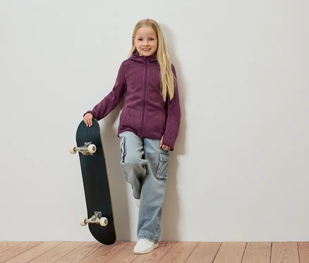 Une fille blonde s'appuie contre un mur, tient un skateboard et porte une veste en polaire violette, un jean et des chaussures blanches.
