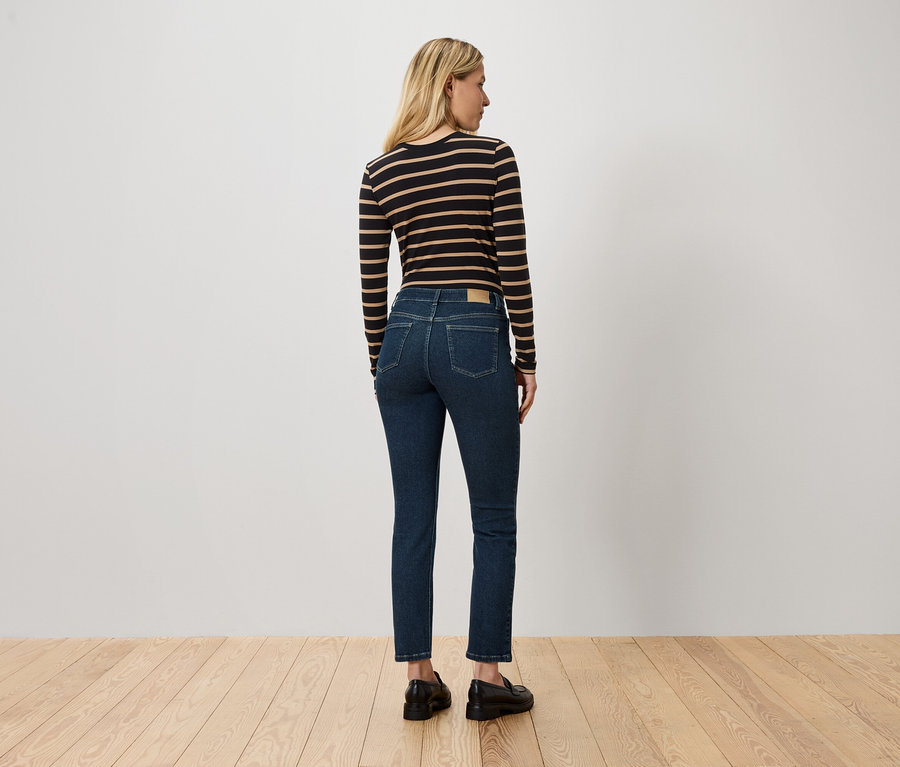 Une femme aux cheveux blonds se tient dos à la caméra, portant un t-shirt à manches longues rayé, un jean bleu et des chaussures noires.