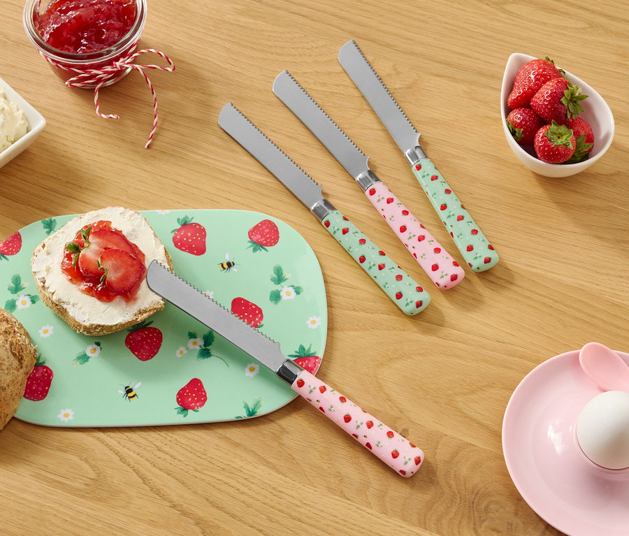 Sur une table en bois se trouvent un pain, une assiette à motifs de fraises, du fromage frais, de la confiture, des fraises et quatre couteaux à motifs de fraises.