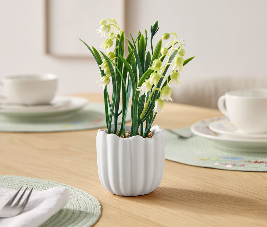 Sur une table, un pot de muguet et à côté des couverts avec des tasses.