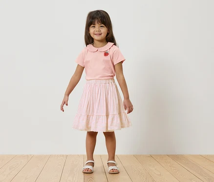 Une jeune fille se tient devant un mur blanc, elle porte un t-shirt rose avec une fraise et une jupe rayée.