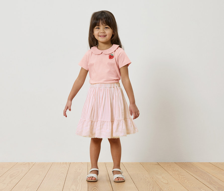 Une jeune fille se tient devant un mur blanc, elle porte un t-shirt rose avec une fraise et une jupe rayée.