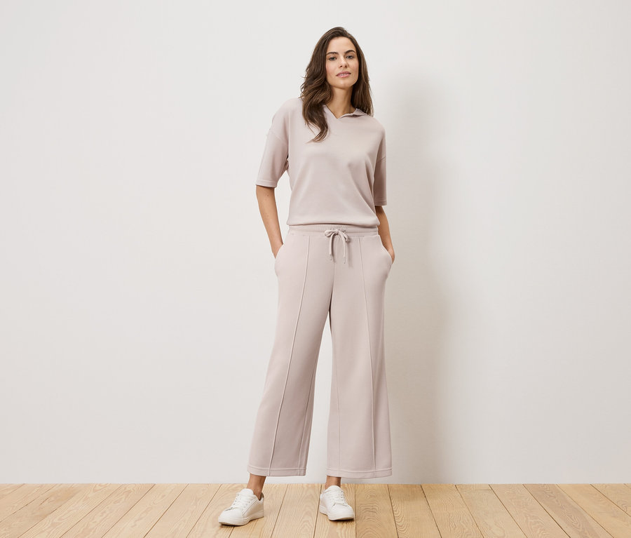Femme portant un tailleur pantalon beige et des baskets blanches debout devant un mur blanc.