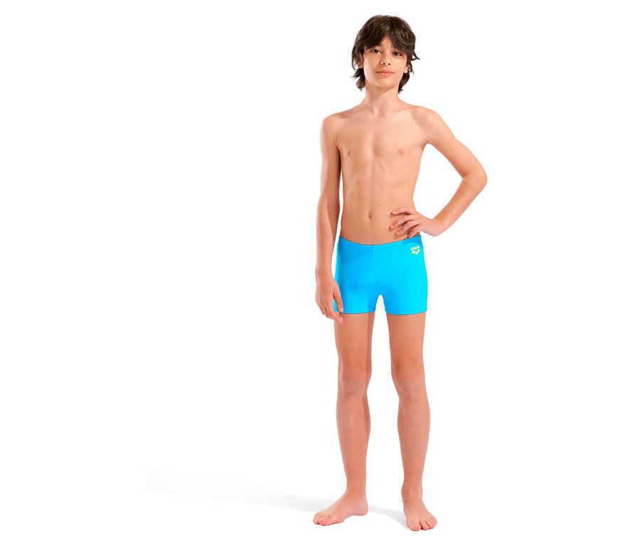 Un jeune homme se tient pieds nus devant un fond blanc et porte un slip de bain bleu.