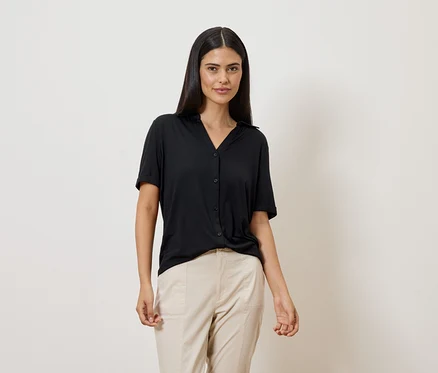Une femme aux longs cheveux foncés porte un chemisier noir à manches courtes et un pantalon beige sur fond blanc.