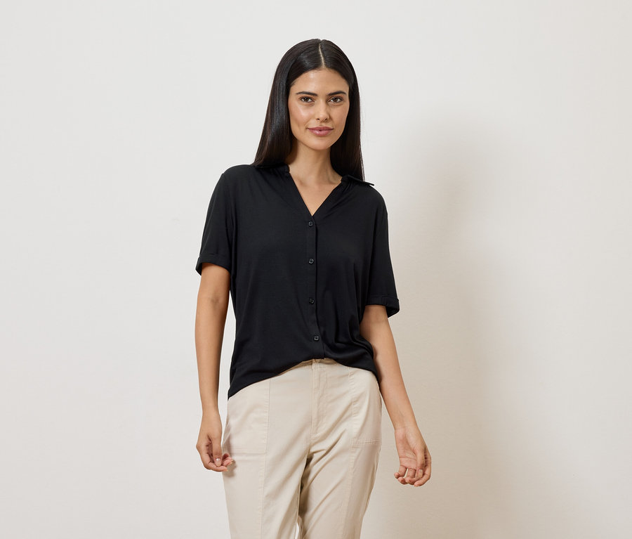 Une femme aux longs cheveux foncés porte un chemisier noir à manches courtes et un pantalon beige sur fond blanc.