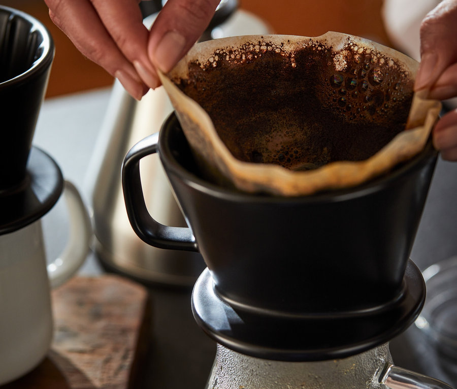 Une femme place un filtre en papier dans un filtre à café manuel noir. En dessous se trouve une cafetière en verre.