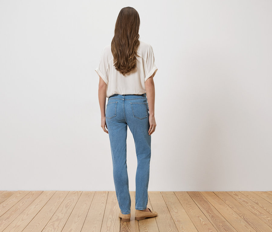 Femme aux longs cheveux bruns, vue de dos, portant un chemisier clair et un jean bleu, debout sur un plancher en bois devant un mur blanc.