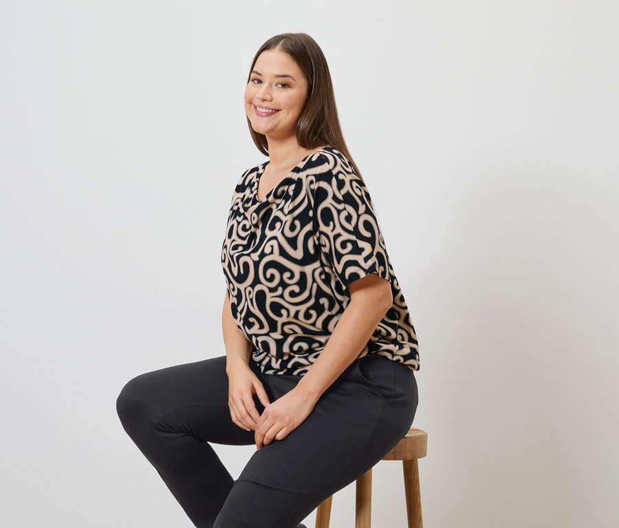 Une femme est assise sur un tabouret, portant un chemisier à motifs et un pantalon de jogging.