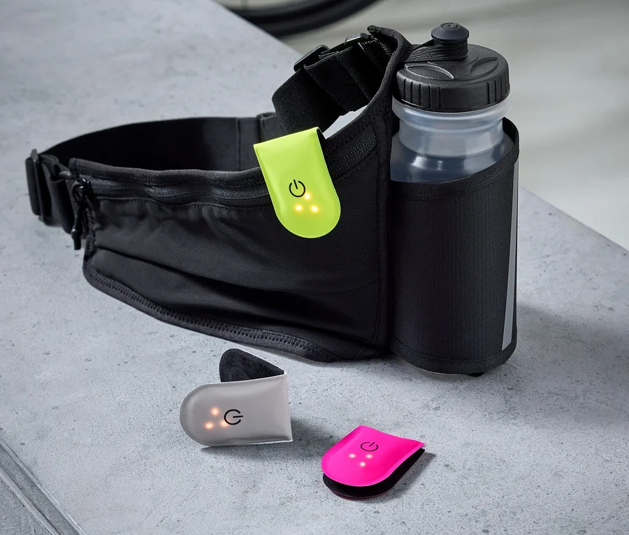 Sac banane noir avec gourde et trois clips lumineux colorés sur fond gris, avec un vélo en arrière-plan.