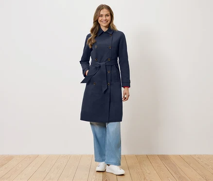Une femme se tient devant un mur blanc, portant un trench-coat bleu marine avec une ceinture, un jean bleu et des baskets blanches.