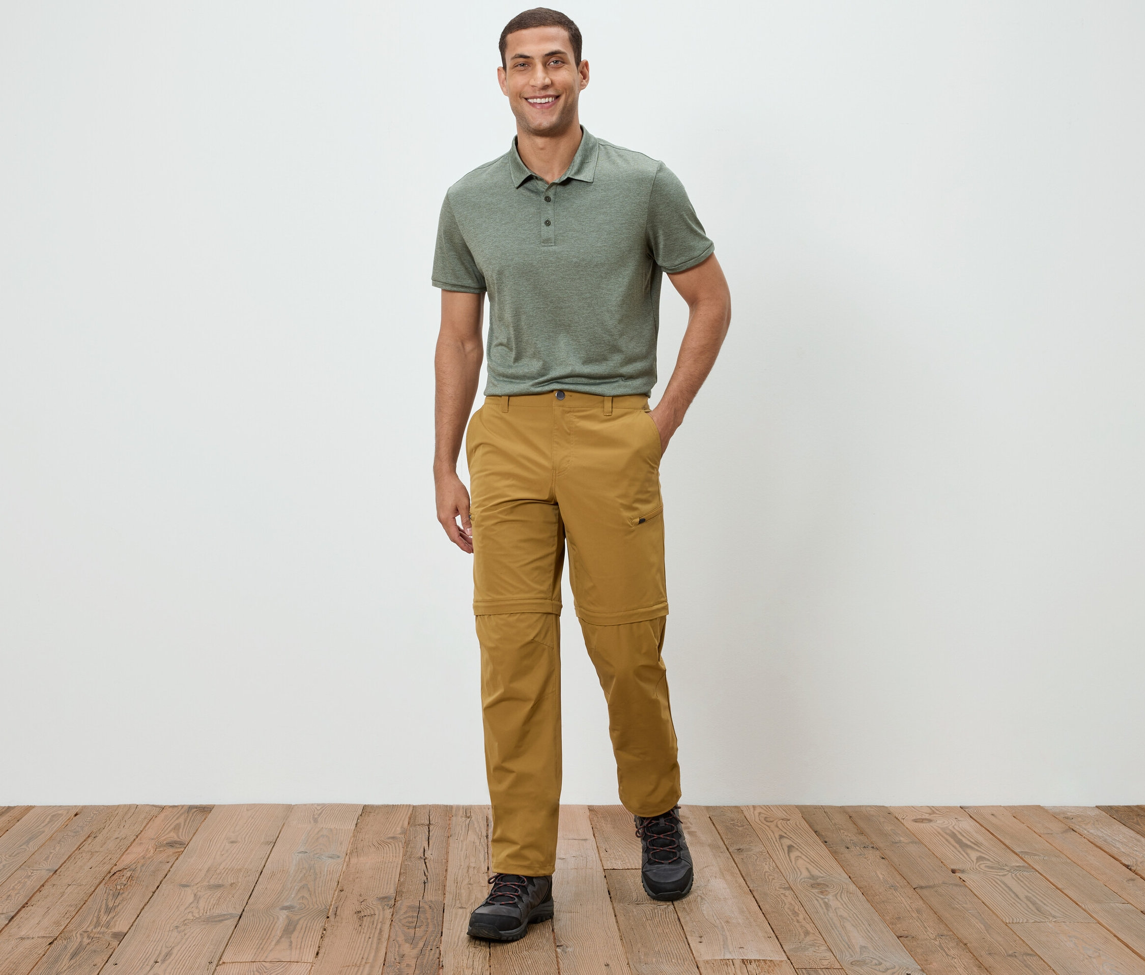 Un homme se tient devant un mur blanc, portant un polo vert et un pantalon kaki.