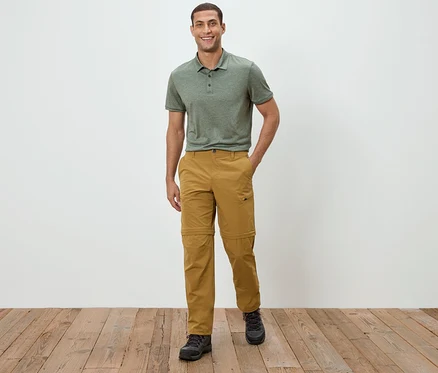 Un homme se tient devant un mur blanc, portant un polo vert et un pantalon kaki.