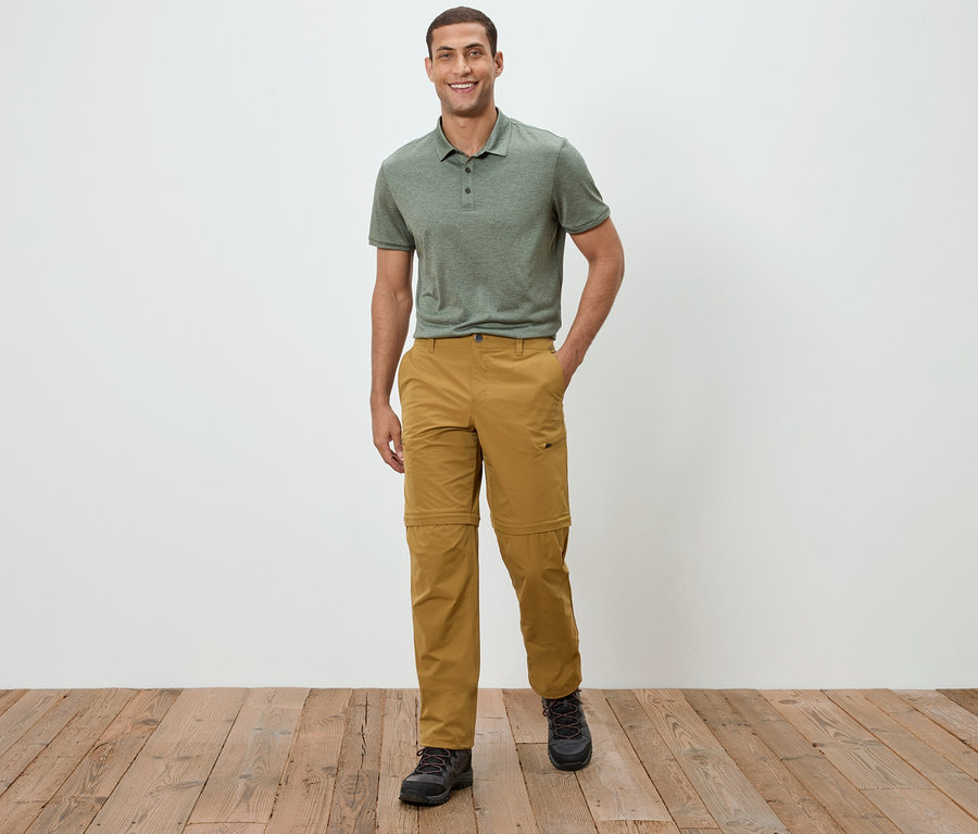 Un homme se tient devant un mur blanc, portant un polo vert et un pantalon kaki.