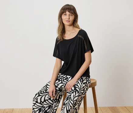Femme assise sur un tabouret, portant un t-shirt noir avec des détails en dentelle et un pantalon à motifs noir et blanc.