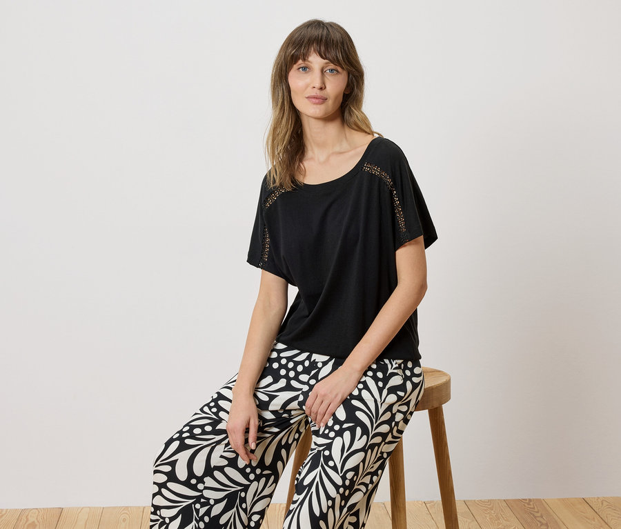 Femme assise sur un tabouret, portant un t-shirt noir avec des détails en dentelle et un pantalon à motifs noir et blanc.