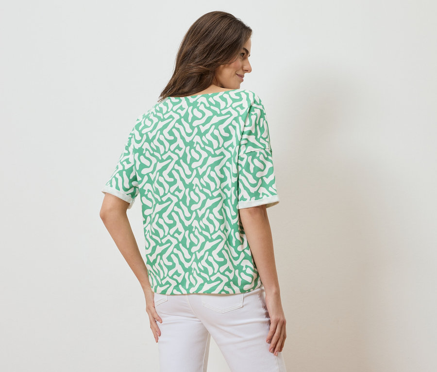 Vue arrière d'une femme aux cheveux bruns portant un haut à manches courtes à motifs verts et blancs et un jean blanc.