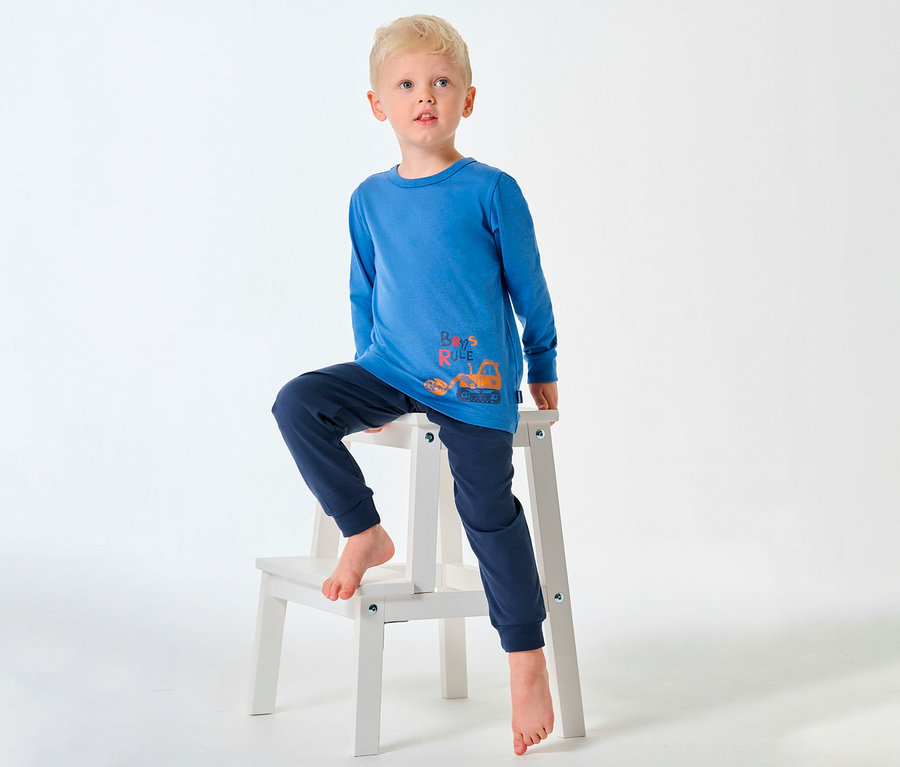 Un garçon blond en pyjama long pour enfant SCHIESSER est assis pieds nus sur un tabouret.