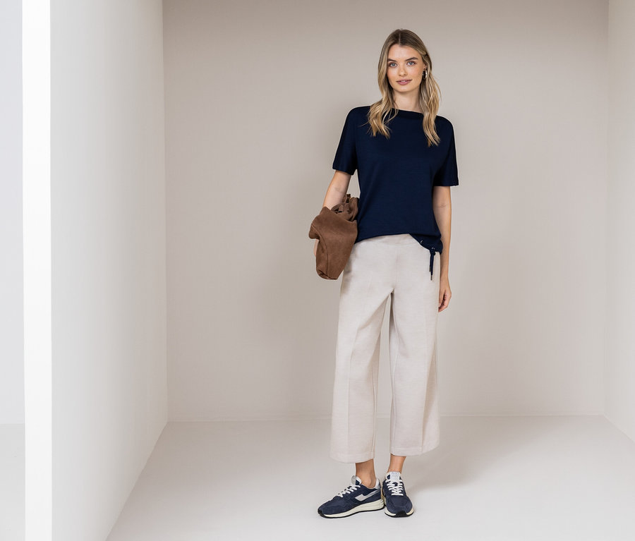 Une femme se tient dans une pièce, portant un t-shirt bleu marine, un pantalon beige et des baskets bleues. Elle tient un sac marron.
