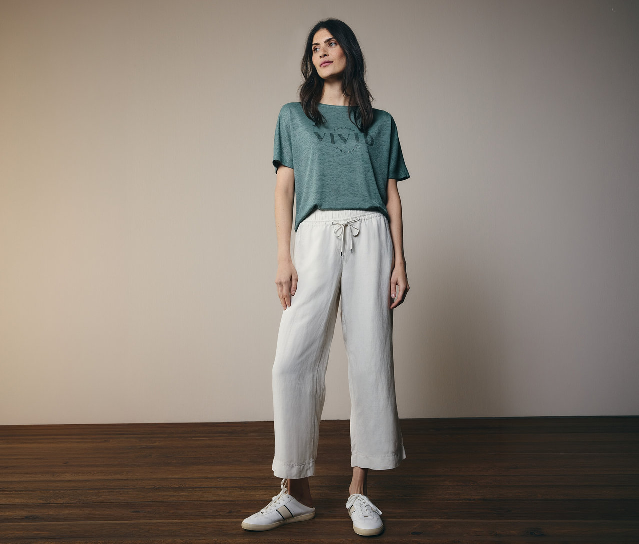Une femme aux cheveux brun foncé porte un t-shirt vert avec l'inscription "VIVID", un pantalon blanc à cordon et des chaussures blanches.