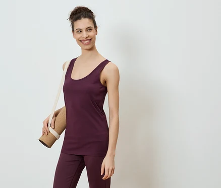 Une femme souriante portant un débardeur et un pantalon aubergine, un tapis de yoga sur l'épaule, sur fond blanc.