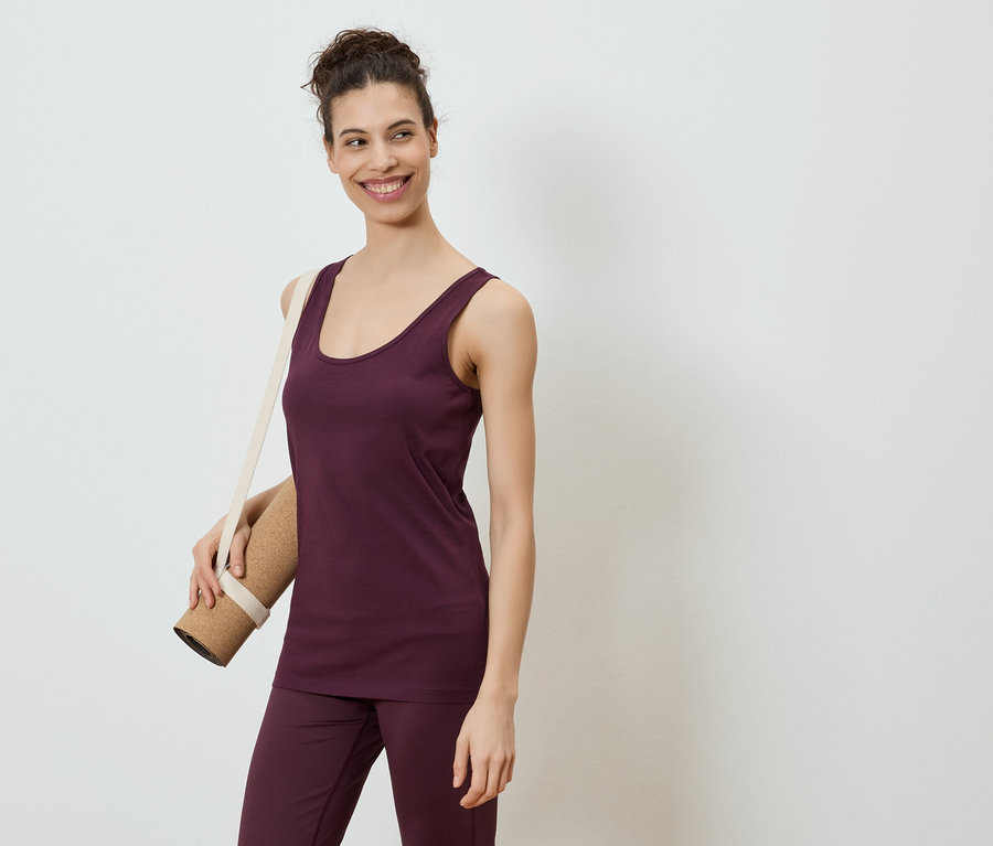 Une femme souriante portant un débardeur et un pantalon aubergine, un tapis de yoga sur l'épaule, sur fond blanc.