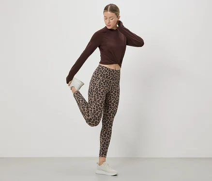 Une femme étire sa jambe en portant un haut marron, un legging à motif léopard et des baskets blanches sur fond blanc.
