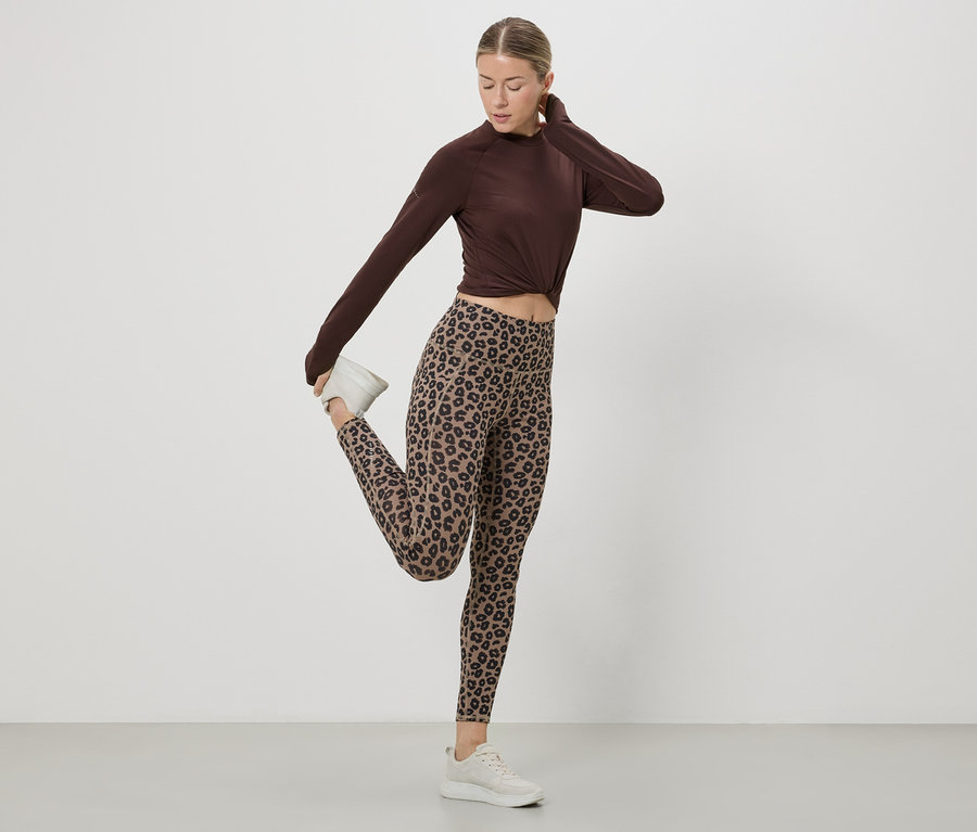 Une femme étire sa jambe en portant un haut marron, un legging à motif léopard et des baskets blanches sur fond blanc.