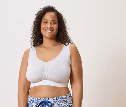 Une femme souriante aux cheveux bouclés porte un soutien-gorge blanc et un short à motifs bleus sur un fond clair.