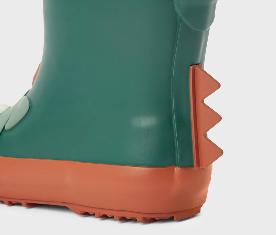 Gros plan d'une botte en caoutchouc verte avec des détails orange et des pointes de dinosaure sur un fond gris clair.