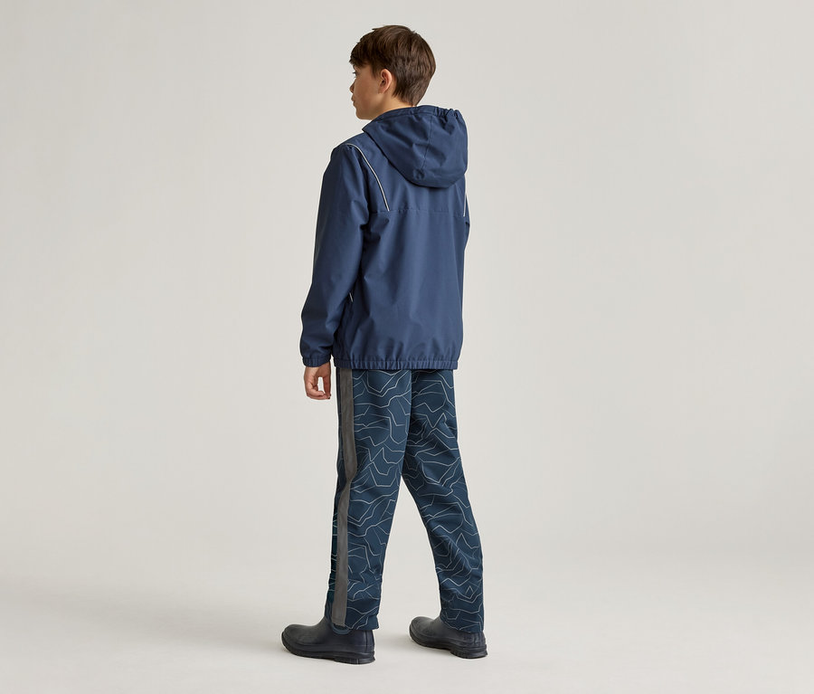 Un garçon est debout, dos à la caméra, portant une veste à capuche bleu foncé avec des rayures blanches, un pantalon bleu foncé à motifs clairs et des bottes sombres.