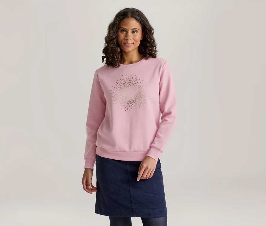 Une femme aux cheveux foncés et bouclés sourit légèrement, vêtue d'un sweat-shirt rose clair à manches longues avec un motif circulaire d'étoiles sur le devant, ainsi que d'une jupe bleu foncé et de collants bleu foncé.