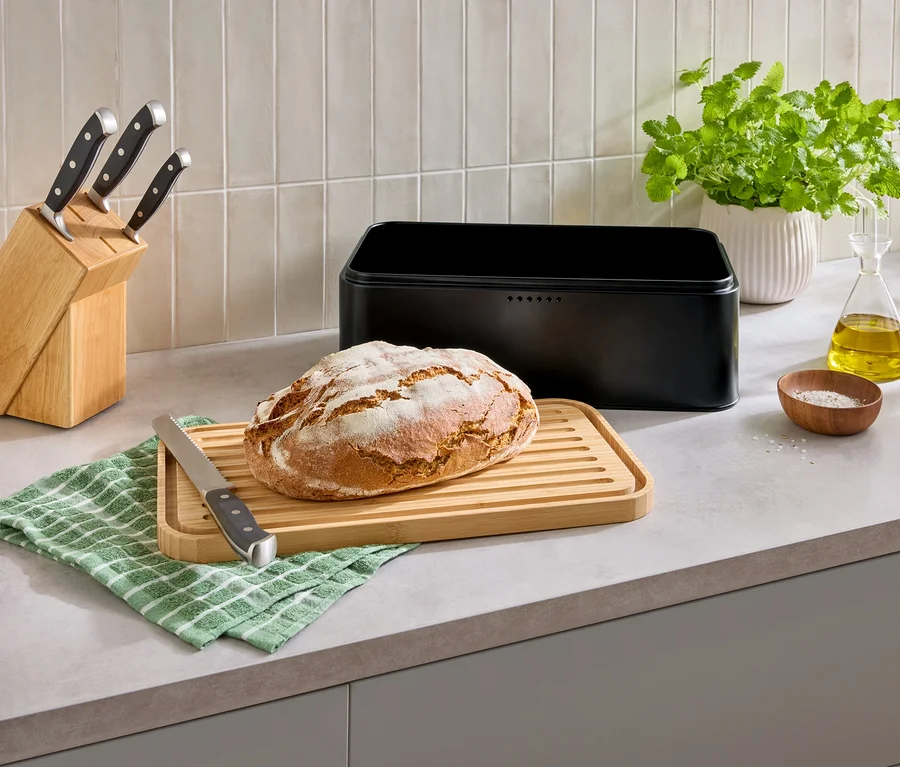 Sur un comptoir de cuisine, un pain sur une planche à découper, une boîte à pain, un bloc de couteaux et des épices.