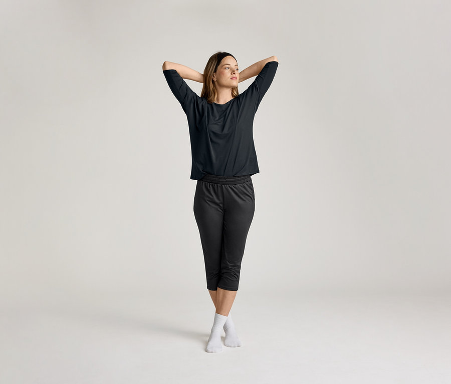 Une femme, les mains croisées derrière la tête, porte un t-shirt de sport 3/4 noir.