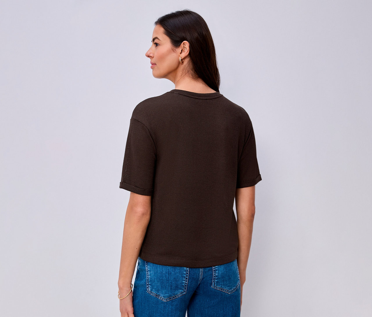 Une femme vue de dos, légèrement tournée vers la droite, porte un T-shirt Street One marron foncé à manches courtes et un jean bleu. Elle a de longs cheveux foncés, de petites boucles d'oreilles dorées et un bracelet doré au poignet gauche.