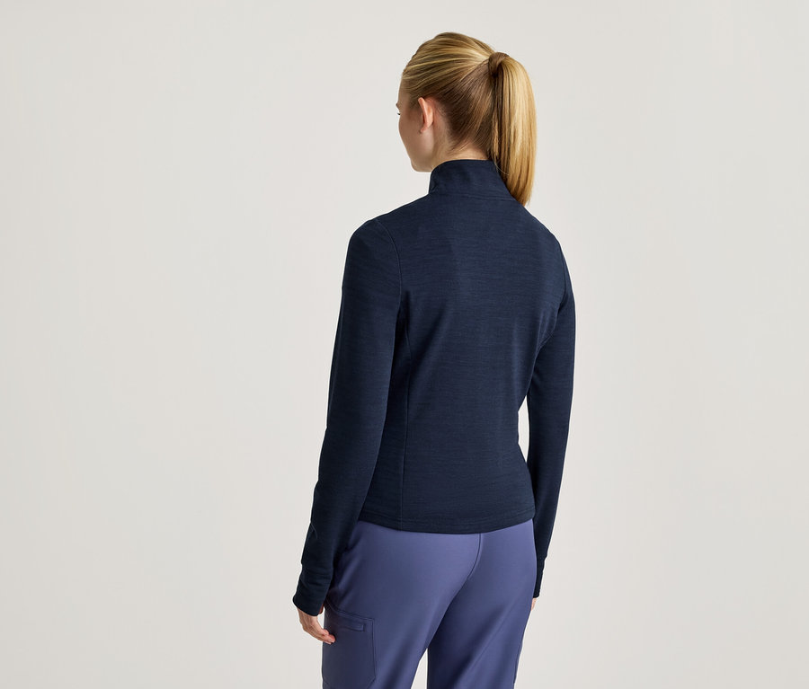 Vue de dos, une femme avec une queue de cheval blonde porte une veste zippée bleu marine et un pantalon bleu.