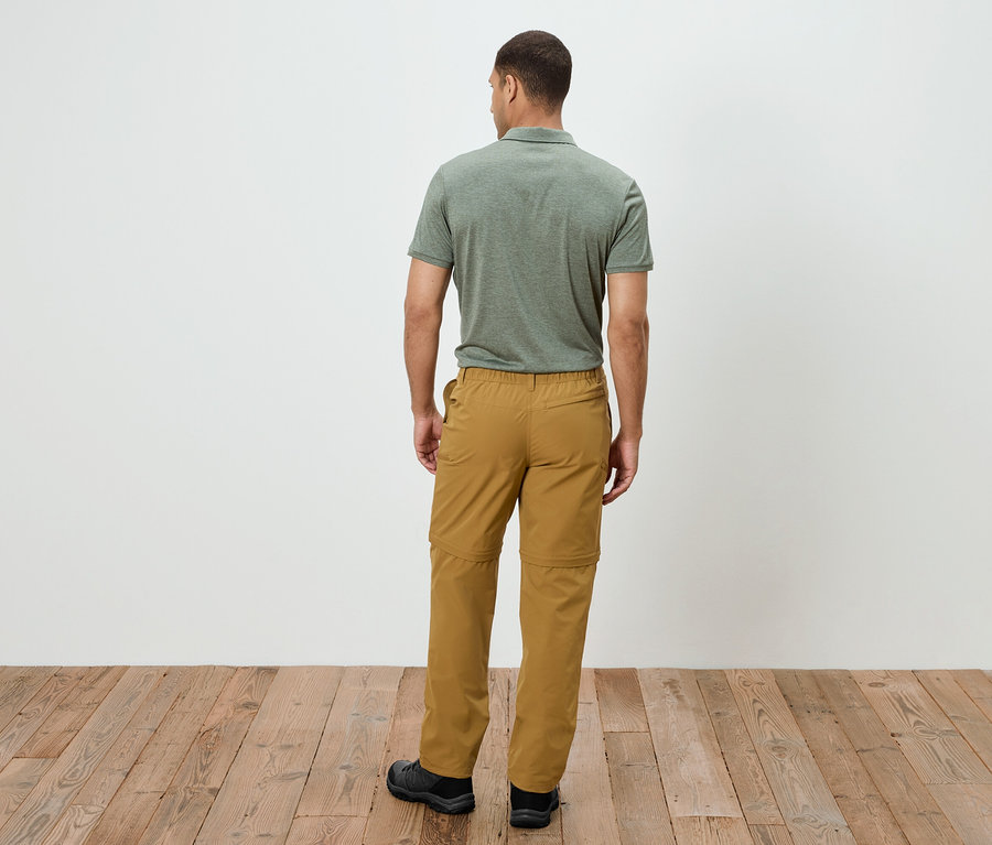 Un homme se tient dos à la caméra, portant un polo vert olive et un pantalon d'extérieur doré avec fermeture éclair. Il se tient sur un plancher en bois.