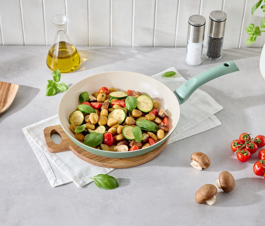 Des gnocchis aux légumes dans une poêle vert clair sur une assiette en bois. À côté se trouvent des champignons, des tomates, une spatule en bois et une bouteille d'huile.